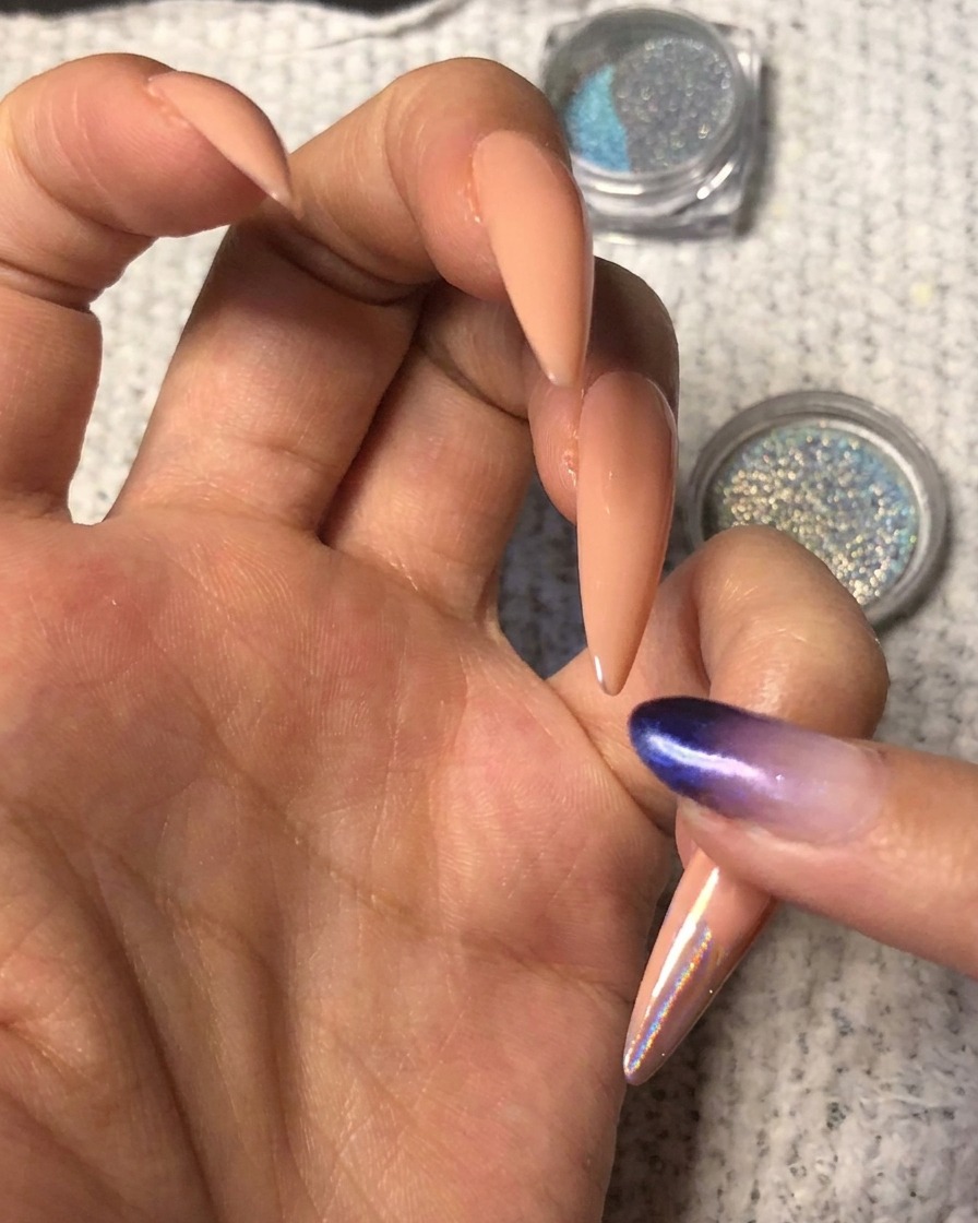 Un dito che preme della vibrante polvere cromata olografica blu-viola sulla punta di ununghia a mandorla nude.