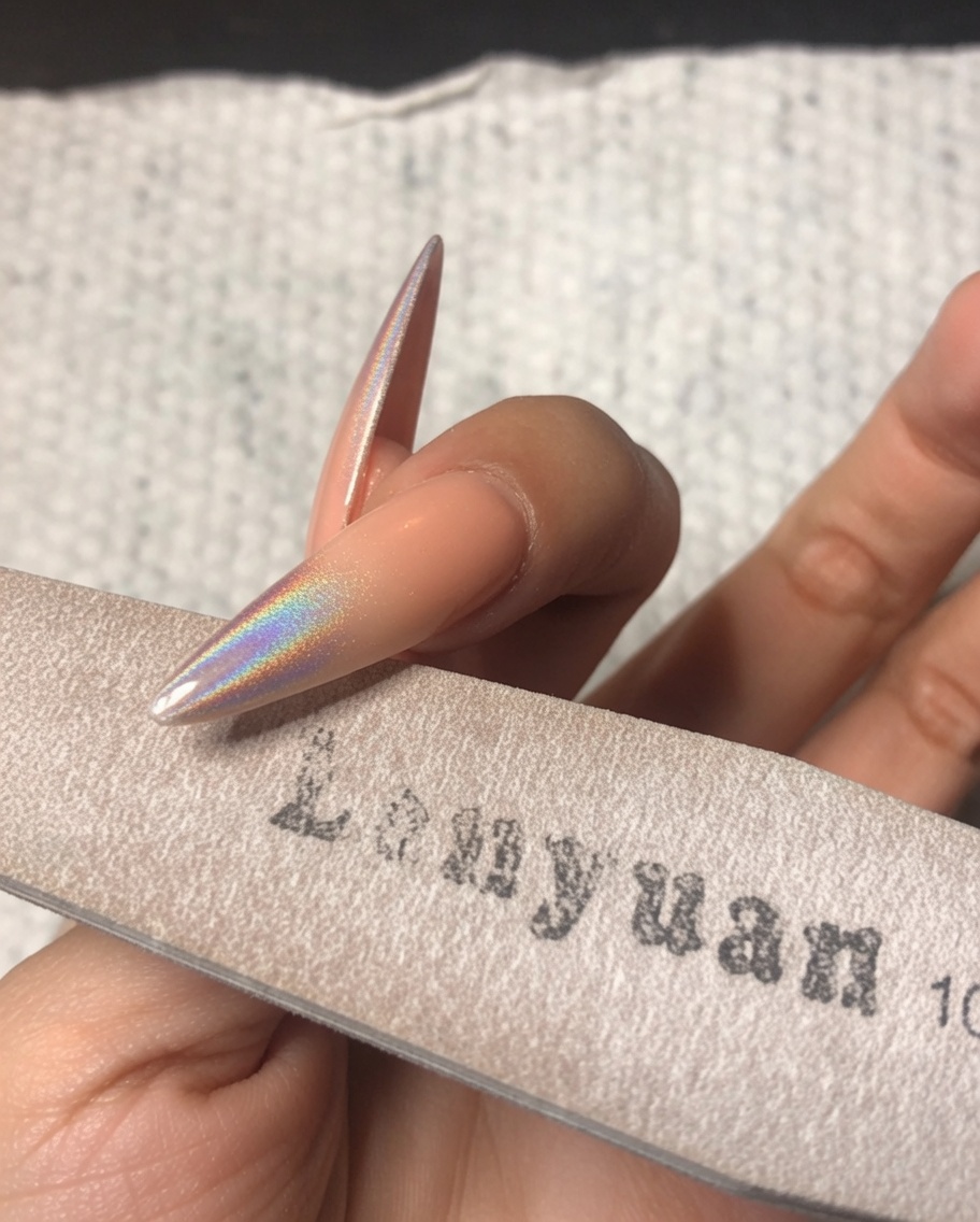 Una mano che usa una lima a spugna fine per levigare delicatamente il bordo libero di ununghia stiletto decorata con polvere cromata olografica.