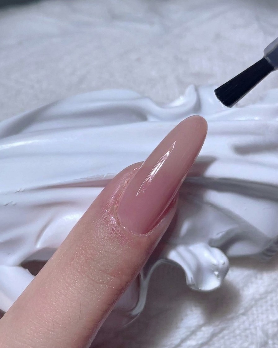 Un pennello per unghie applica un top coat trasparente e lucido su ununghia color nude.