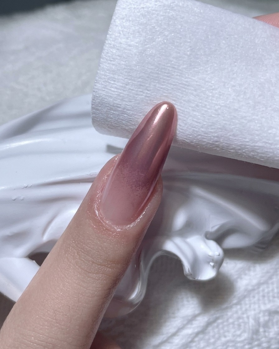 Uno scatto ravvicinato di una mano che usa un pad in cellulosa per pulire i bordi di ununghia con finitura cromata oro rosa.
