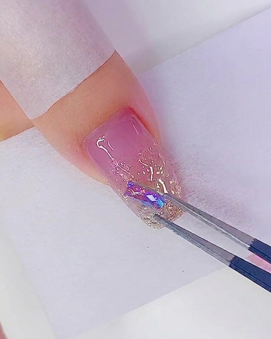 Una pinzetta posiziona uno strass cangiante su un punto di adesivo su ununghia glitterata.