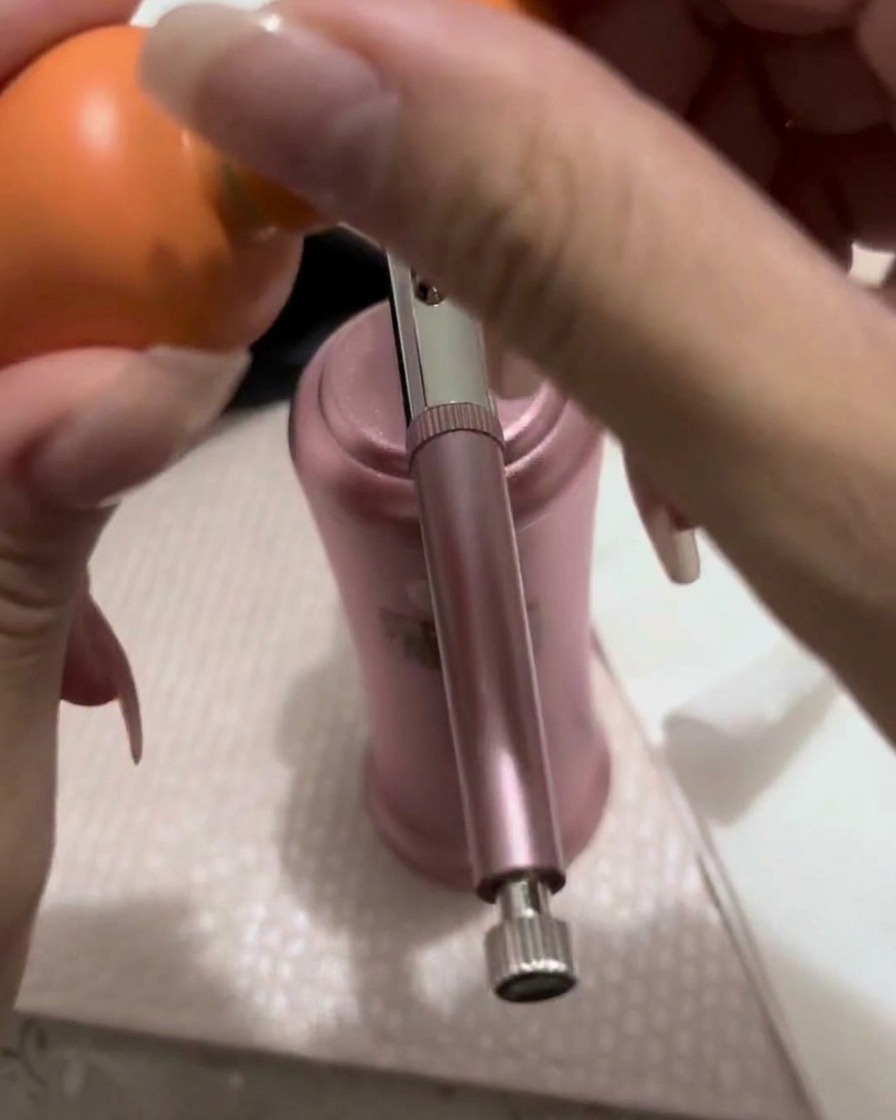 Una piccola quantità di gel color pesca-rosa viene trasferita con un pennello nel serbatoio metallico di una pistola per aerografo.