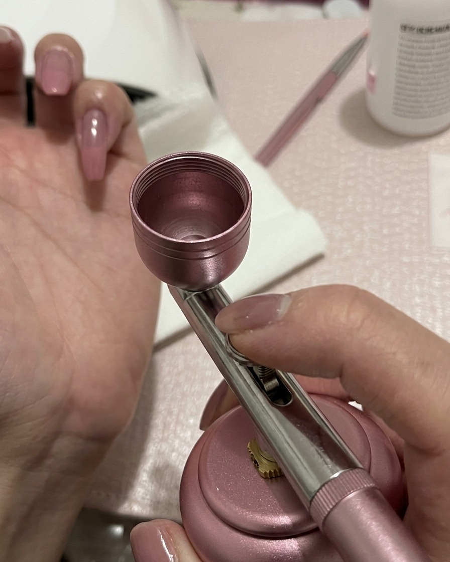 Un aerografo che applica una leggera spolverata di gel pesca-rosa al centro di ununghia preparata.