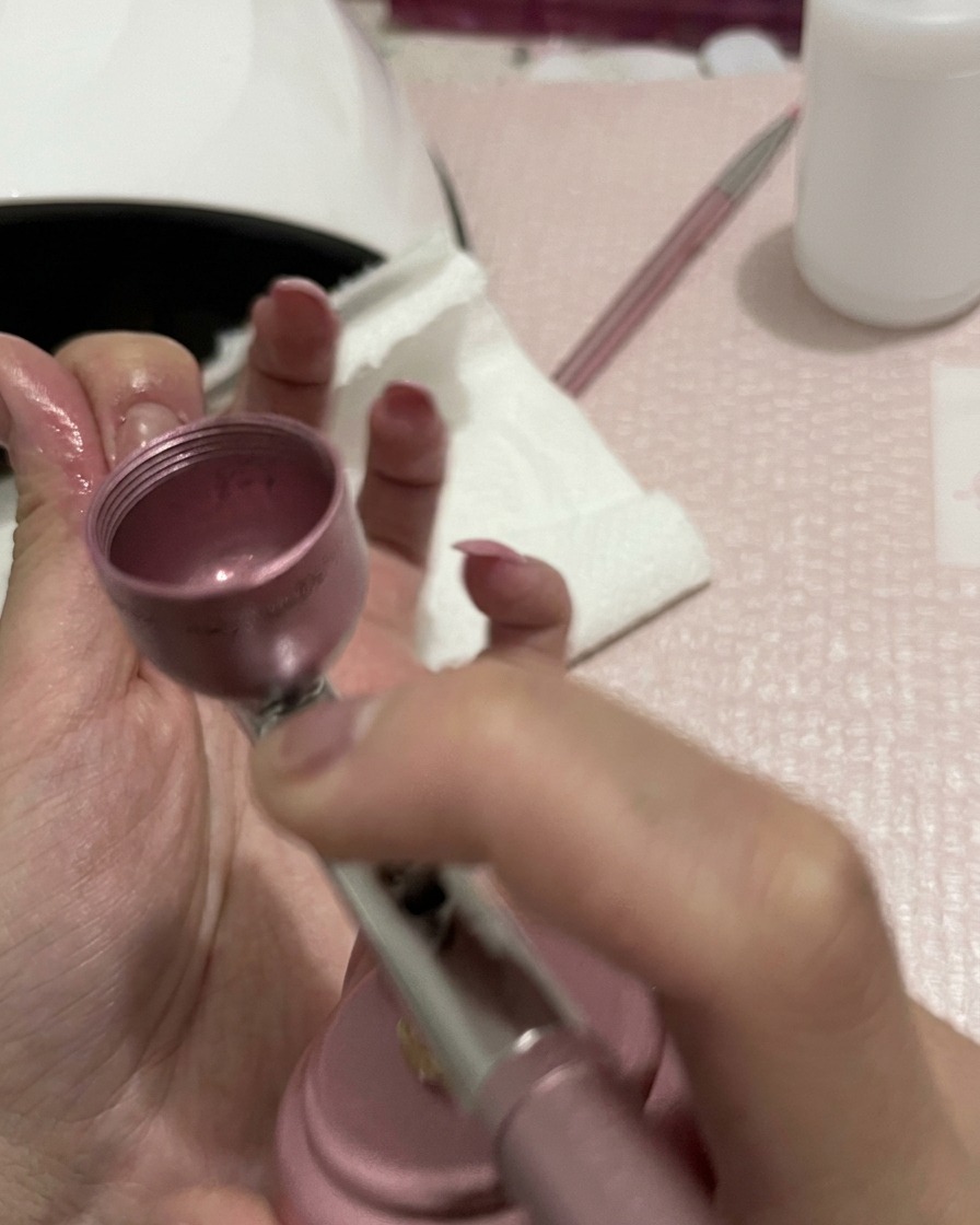 Dettaglio delle mani che tengono un aerografo, con lugello focalizzato sulla sfumatura del colore pesca sulle unghie.