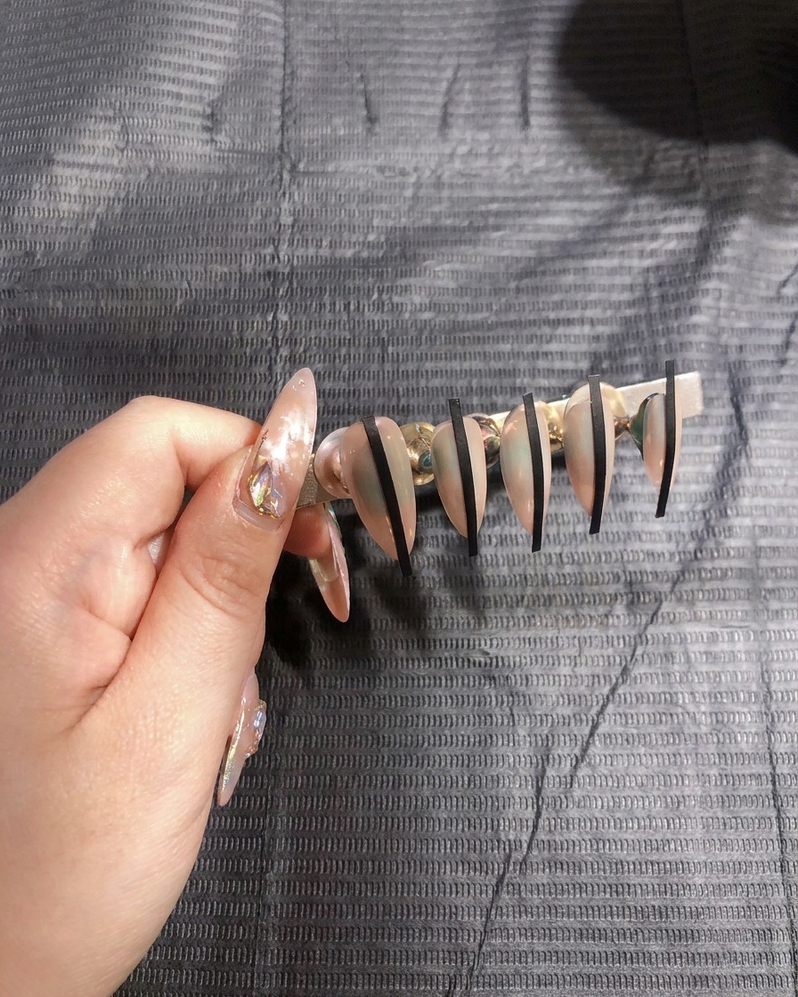 Primo piano di una mano che posiziona un sottile adesivo nero verticalmente su ununghia color nude.
