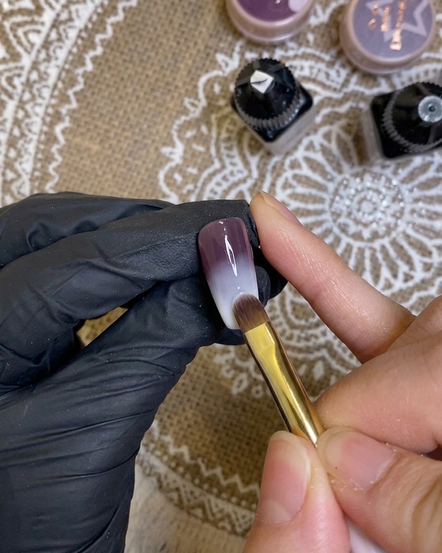 Primo piano di un pennello che applica un gel viola intenso sulla punta di ununghia.