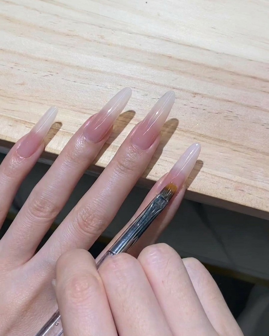 Un pennello da ombré a denti fini che trascina delicatamente verso il basso il gel bianco latte ancora bagnato su una ricostruzione stiletto trasparente.