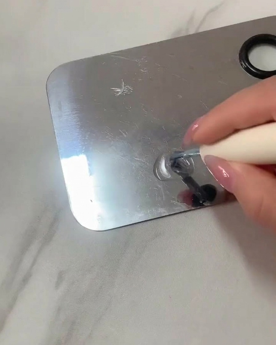 Un pennello piatto in gel mescola gel trasparente e smalto azzurro su una tavolozza da trucco in acciaio inossidabile.