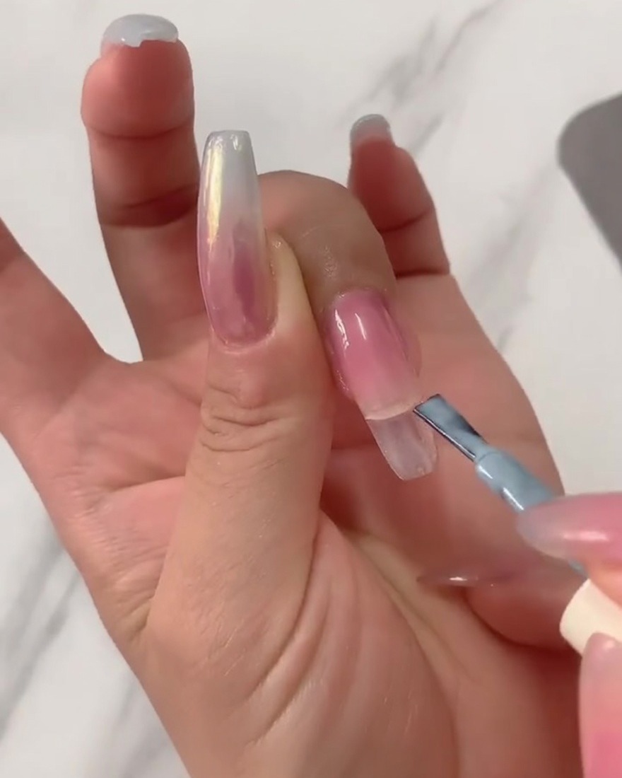 Primo piano di un pennello per gel che applica smalto gel azzurro sulla metà superiore di una tip trasparente lunga a ballerina.
