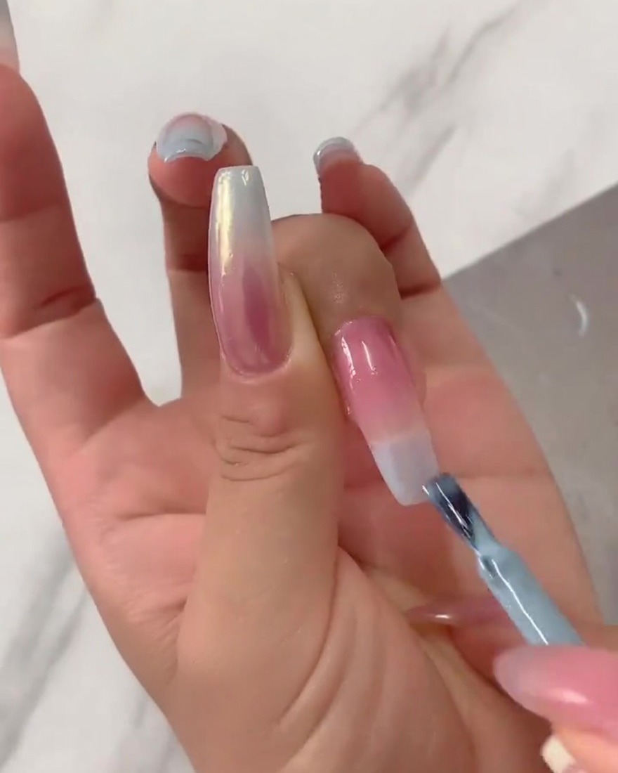 Primo piano di un pennello piatto che sfuma verso il basso lo smalto gel azzurro concentrato dalla punta dellunghia su una base rosa nude velata.
