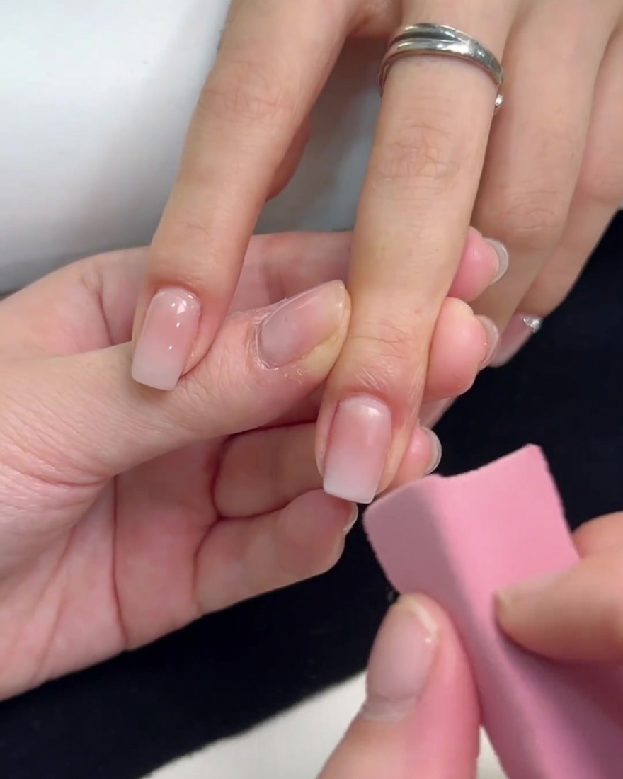 Una spugnetta rosa utilizzata per picchiettare gel bianco sulla punta di ununghia quadrata, sfumandolo verso il basso per creare un effetto ombre.