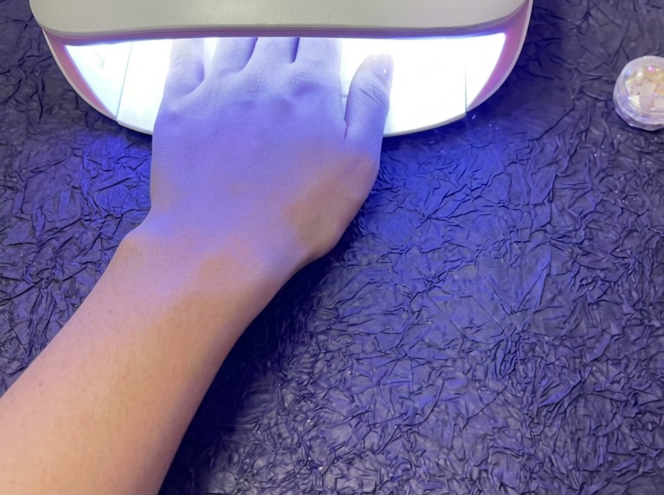 Una mano che riposa allinterno di una lampada per unghie UV/LED accesa per polimerizzare lo smalto gel, poggiata su una superficie viola testurizzata.