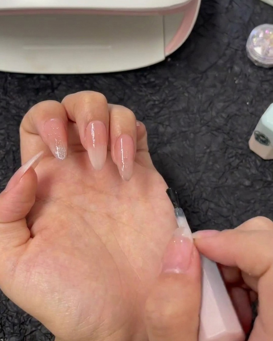 Un pennello che applica il gel di rinforzo trasparente direttamente sullunghia di un indice con base sfumata nude.