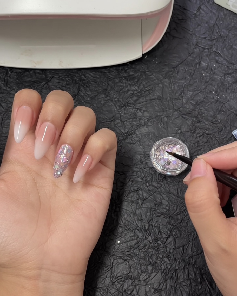 Un pennello per nail art di precisione che si immerge in un piccolo vasetto pieno di scaglie di glitter spesse e iridescenti.