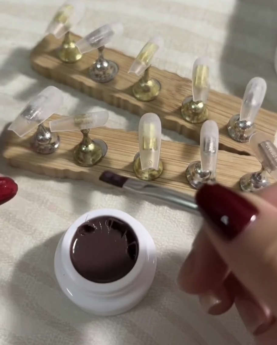 Un pennello piatto che applica uniformemente uno strato velato di smalto gel nude scuro su una tip per unghie press-on trasparente.