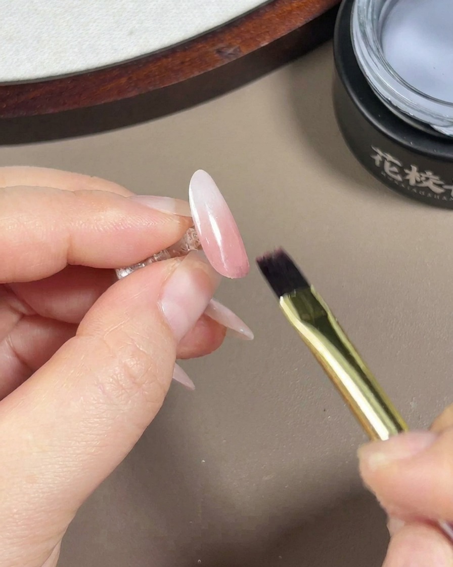 Un pennello per unghie in gel che applica un top coat trasparente e lucido su ununghia finita con sfumatura rosa e bianca e finitura aurora.