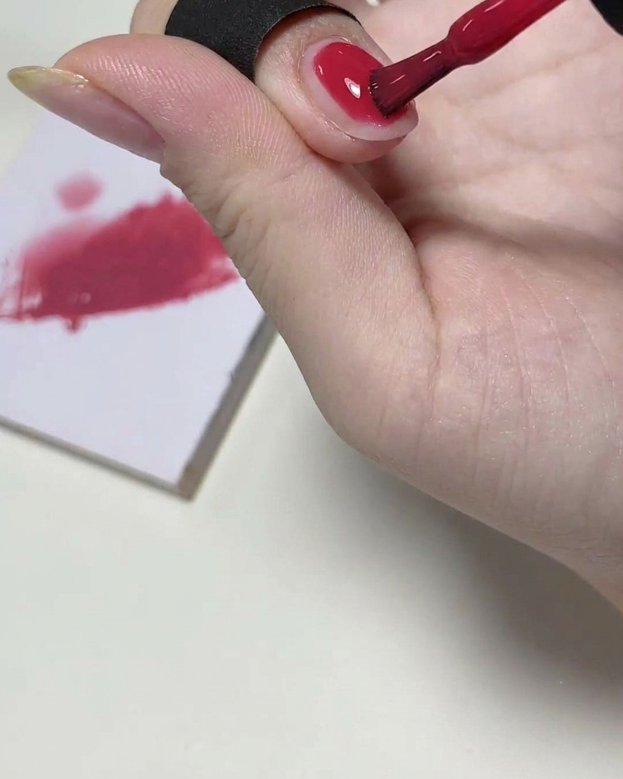 Un pennello per gel applica uno strato leggero di smalto rosa sulla metà superiore dellunghia, con una tavolozza di miscelazione sullo sfondo.