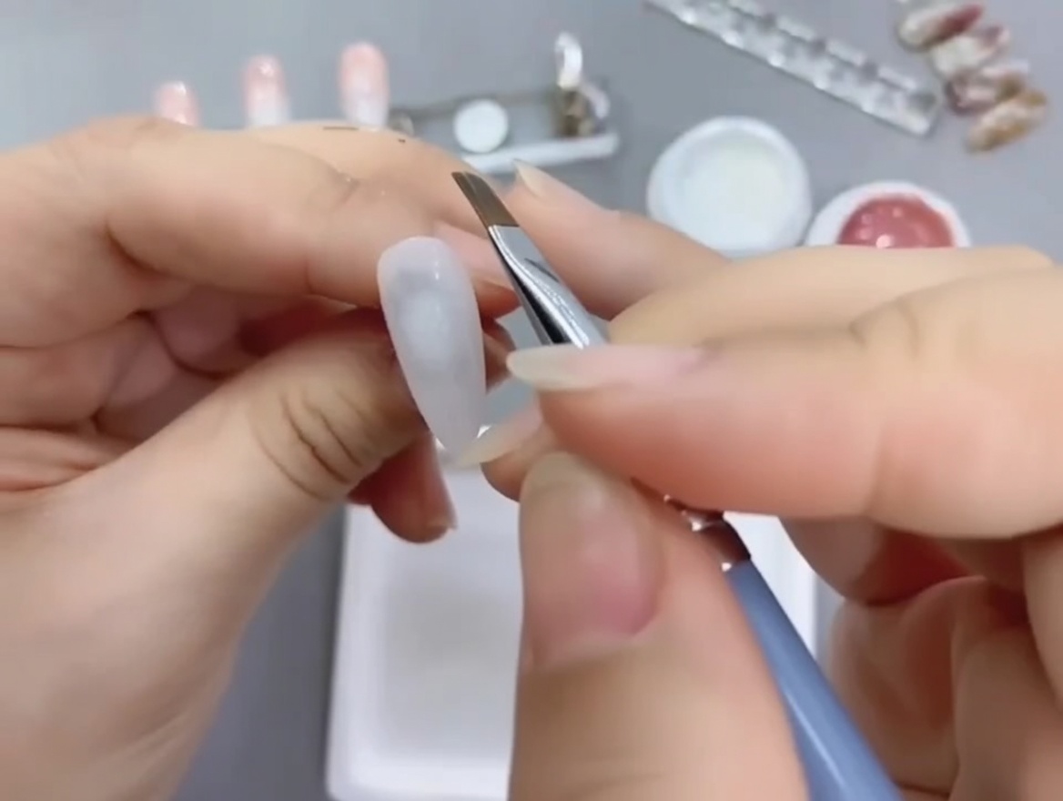 Un pennello angolato per nail art applica gel nude sulla metà inferiore di ununghia base bianca.