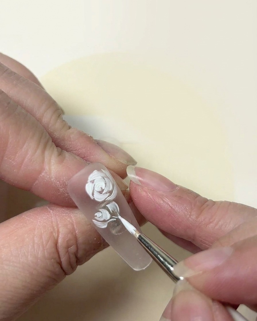 Un primo piano pittorico di un design di rosa bianca rifinito con un pennello a punta fine su ununghia trasparente.