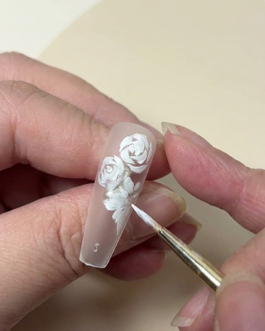Primo piano della punta di ununghia che mostra una rosa bianca dipinta completata da un piccolo fiore a cinque petali e dettagli di foglie.