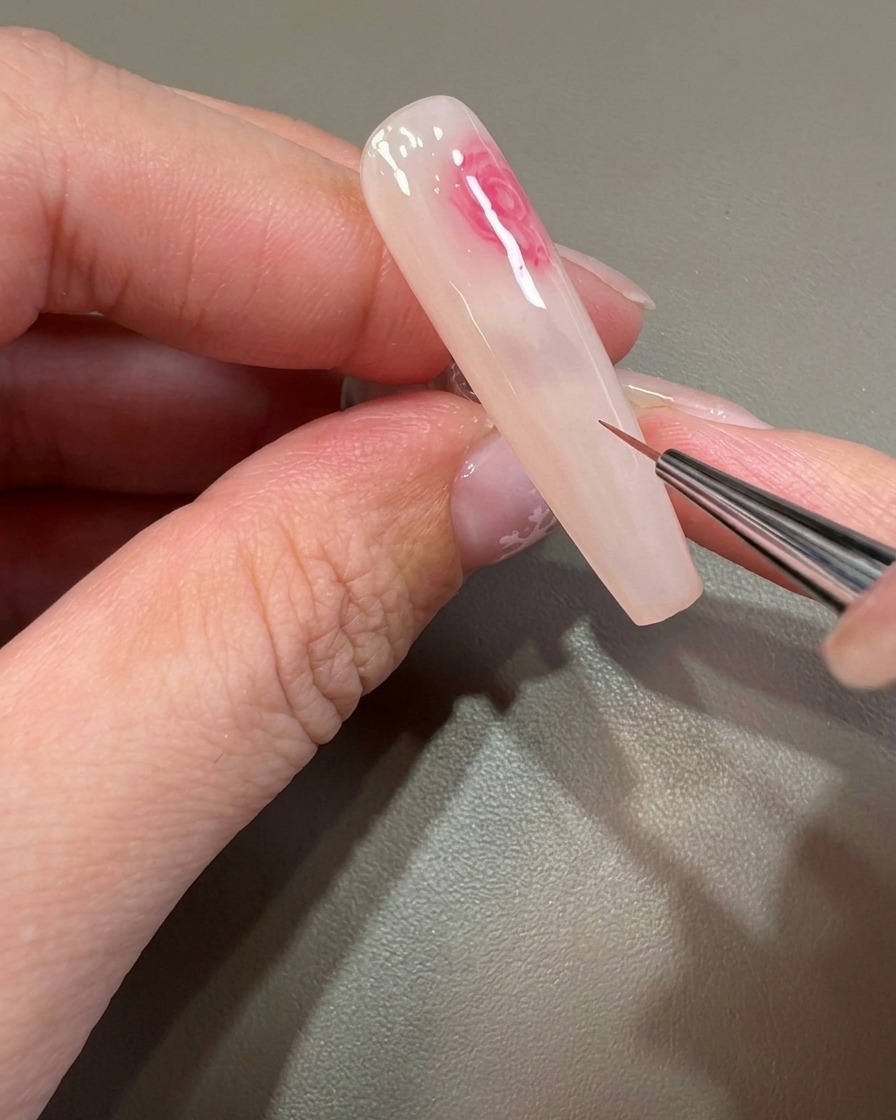Un pennello fine lavora su un pattern floreale dai toni rosa e contorni sfumati sullunghia.