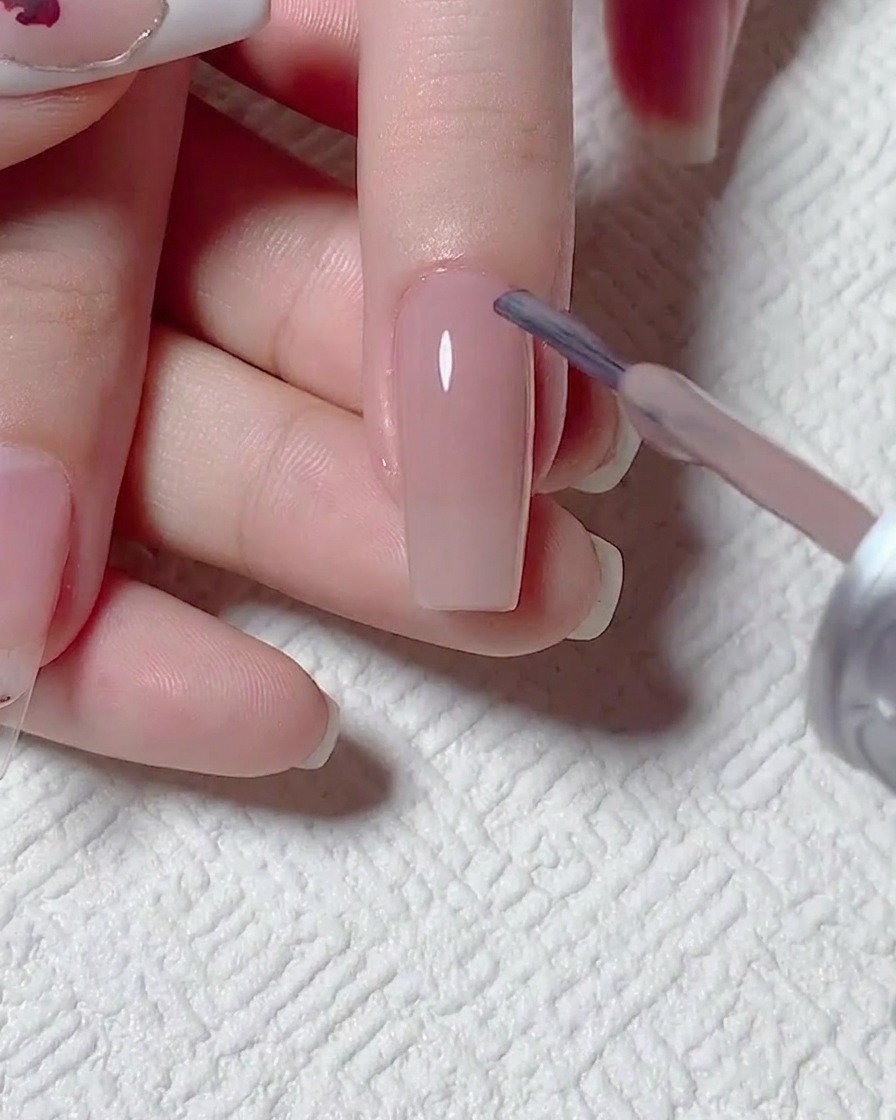 Primo piano di un pennello che applica uno smalto gel nude semitrasparente su una superficie ungueale preparata.