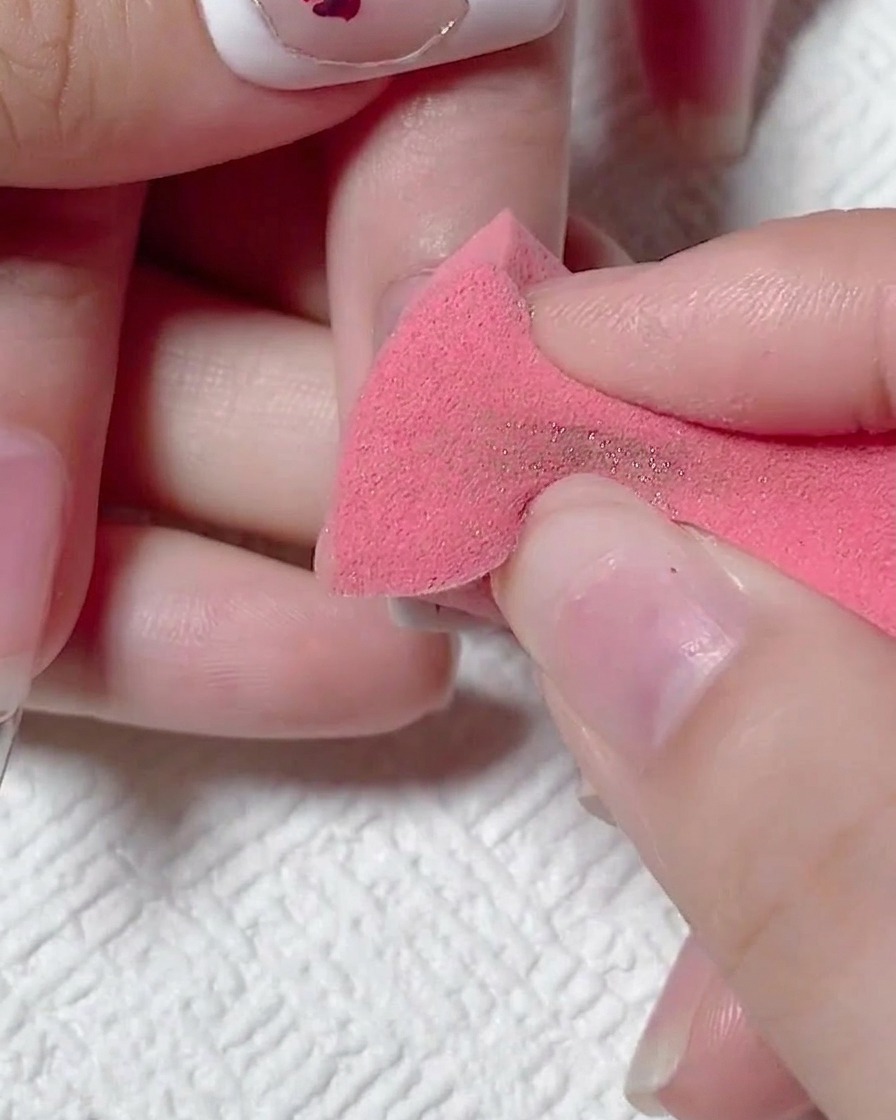 Una mano che usa una spugnetta per trucco rosa per rimuovere la polvere oro in eccesso da ununghia opaca.
