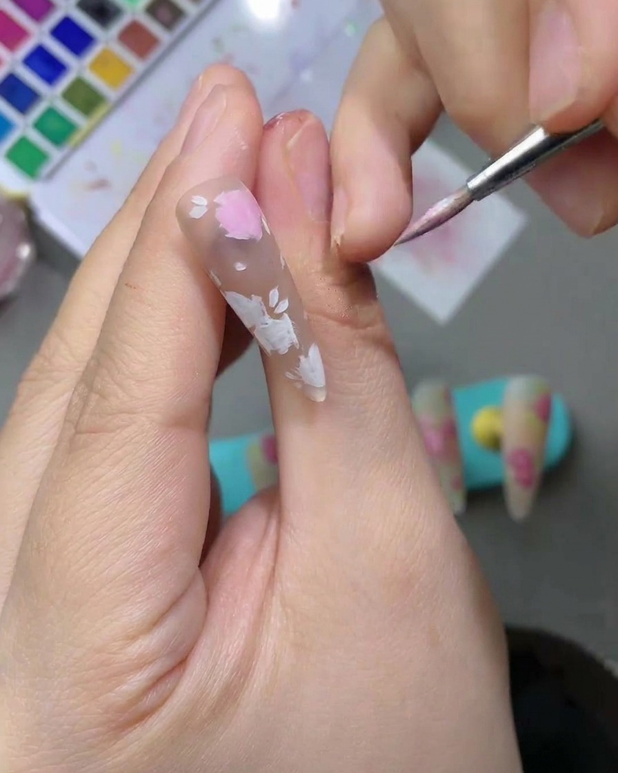 Primo piano di ununghia con delicate forme di petali ad acquerello rosa chiaro che vengono riempite usando un pennello da nail art a punta fine.