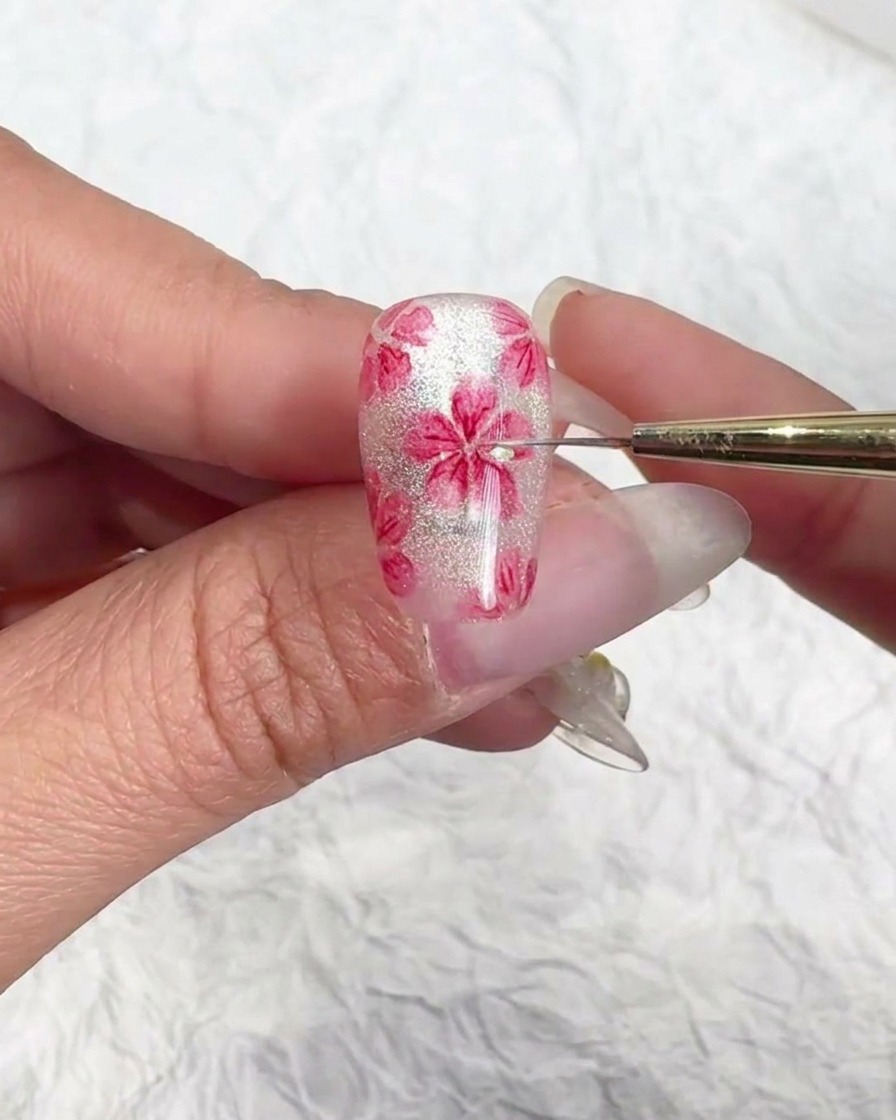 Dettaglio ravvicinato di un pennello per nail art che aggiunge scaglie lucide al centro di un fiore di ciliegio rosa su ununghia cat-eye traslucida.