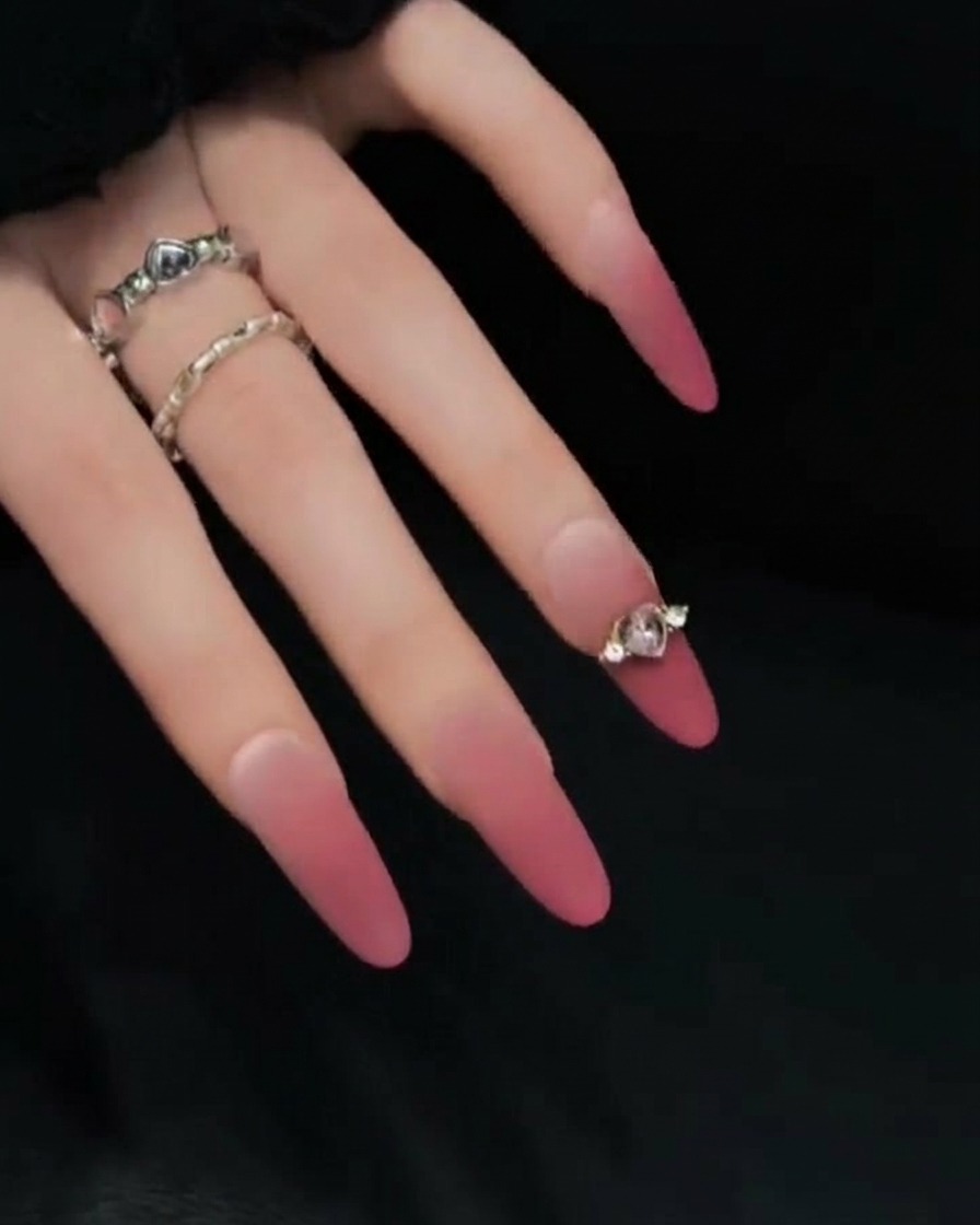 Primo piano di una mano in posa su sfondo scuro, che mostra il profilo laterale di lunghe unghie finte sfumate rosa e nude per evidenziarne lunghezza e curvatura.