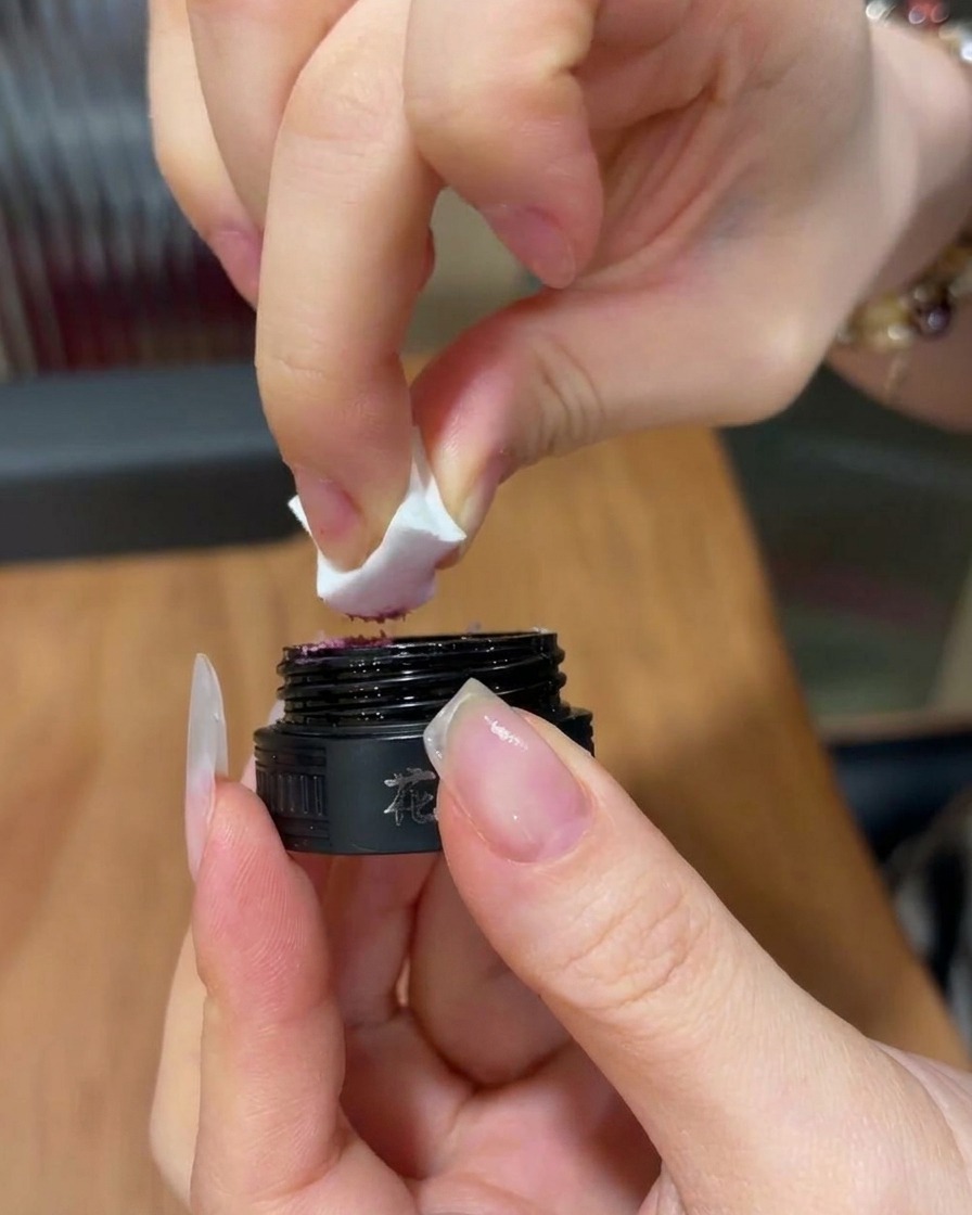 Un primo piano di una mano che tiene una spugnetta per trucco, prelevando una piccola quantità di gel rosa brillante da un vasetto cosmetico nero.