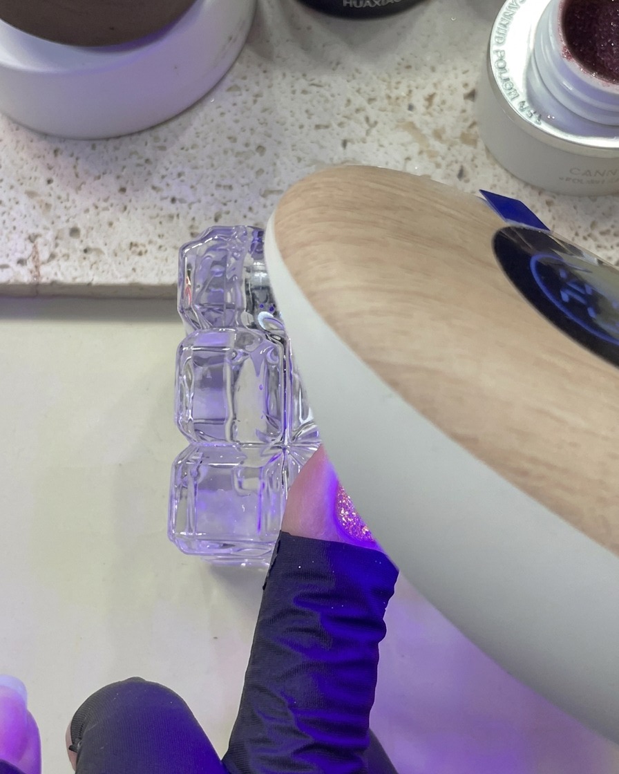 Una mano posizionata sotto una lampada UV/LED mentre si polimerizza ununghia con glitter rosa applicato.