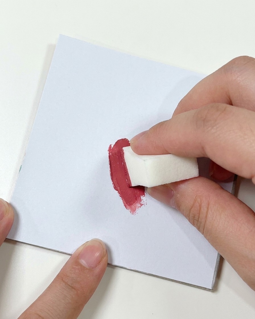 Una mano che preme una piccola spugnetta per il trucco bianca in una macchia di gel rosa scuro su una superficie piatta.
