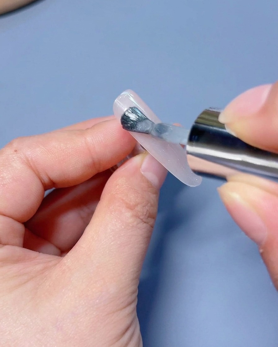 Un pennello che stende uno strato di smalto gel cat-eye bianco scintillante su una tip.