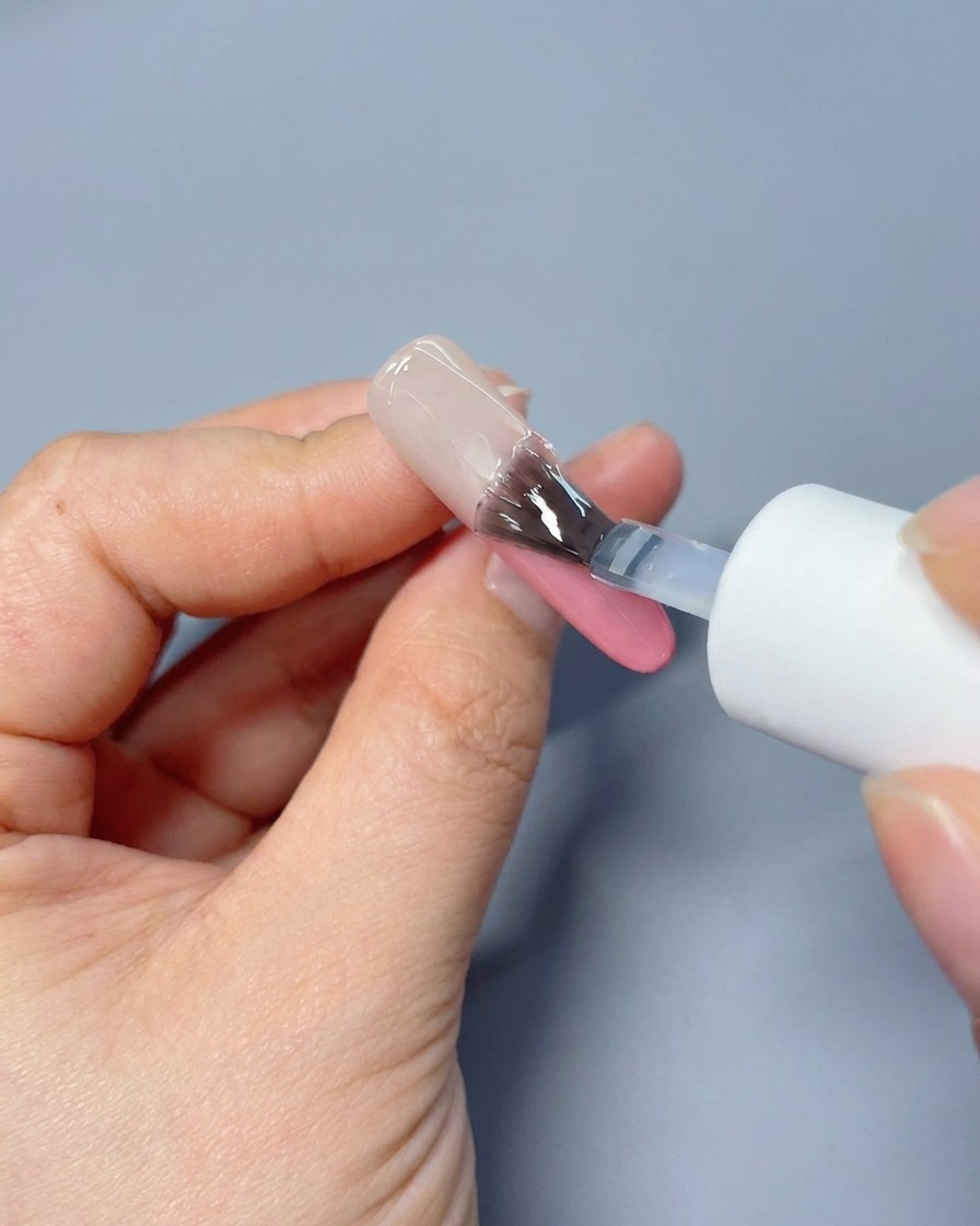 Un pennello che stende un top coat lucido e trasparente su ununghia sfumata rosa.