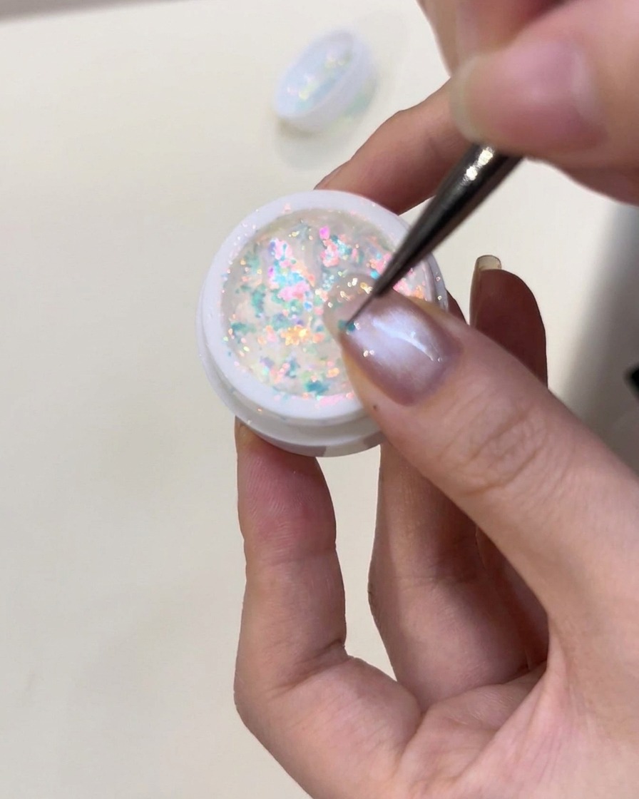 Pinzette che posizionano con cura piccole scaglie iridescenti blu brillanti su ununghia rivestita di gel costruttore trasparente bagnato.