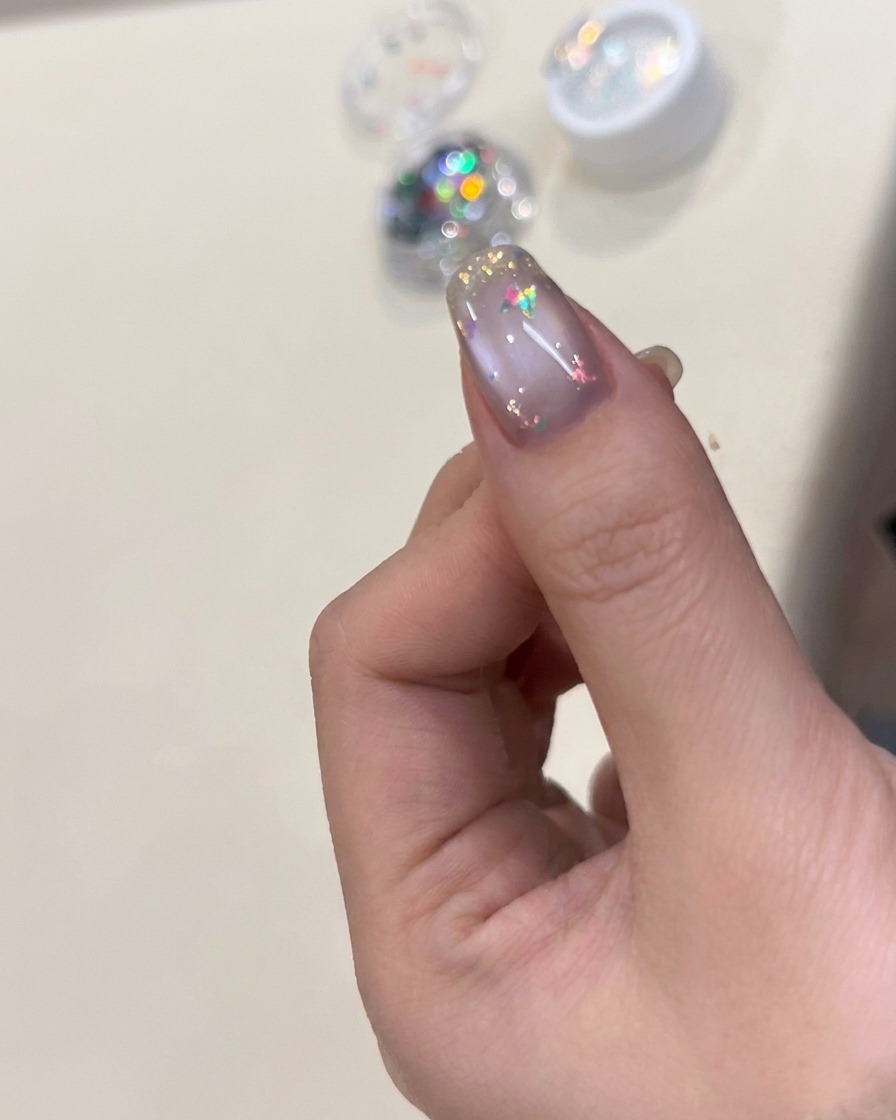 Un pennello per unghie che applica un top coat trasparente e lucido su ununghia decorata con scaglie iridescenti e un charm a farfalla.