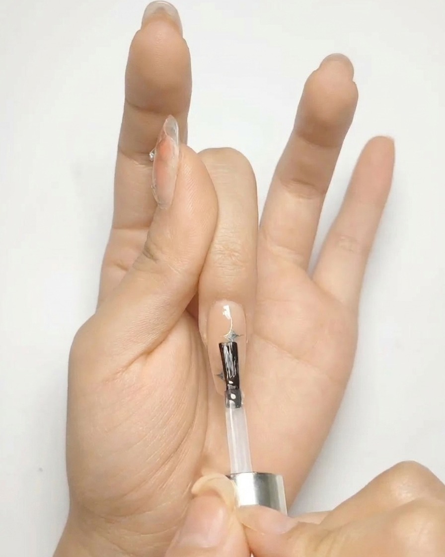 Un pennello applica un top coat trasparente e lucido su ununghia nude decorata con stelle argento.