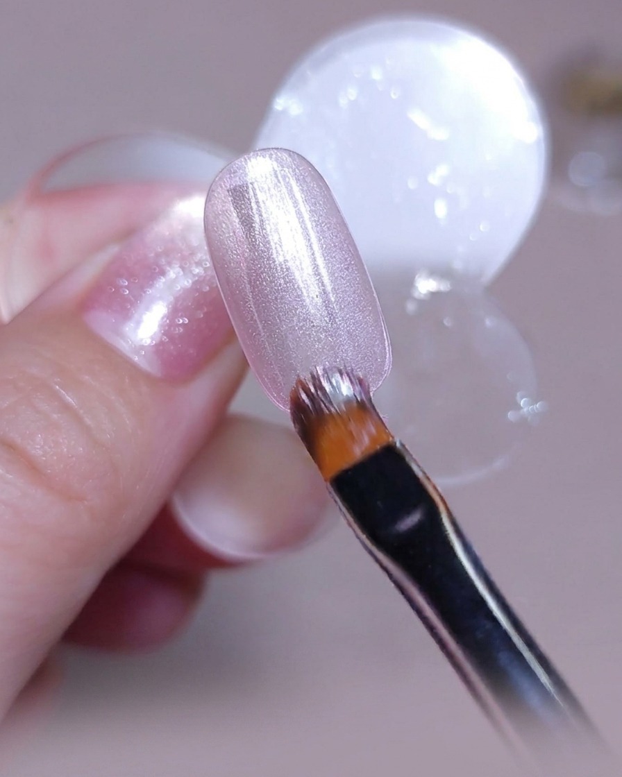 Un pennello applica uno strato uniforme di smalto gel cat-eye argento rosato su una tip per unghie trasparente.