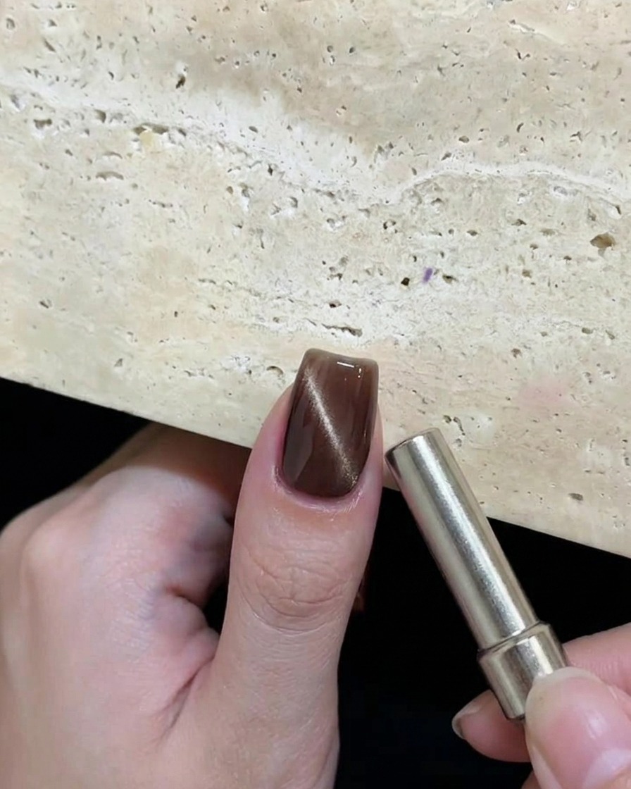 Una mano tiene un magnete cilindrico vicino a ununghia con gel cat-eye marrone per creare un motivo circolare riflettente.