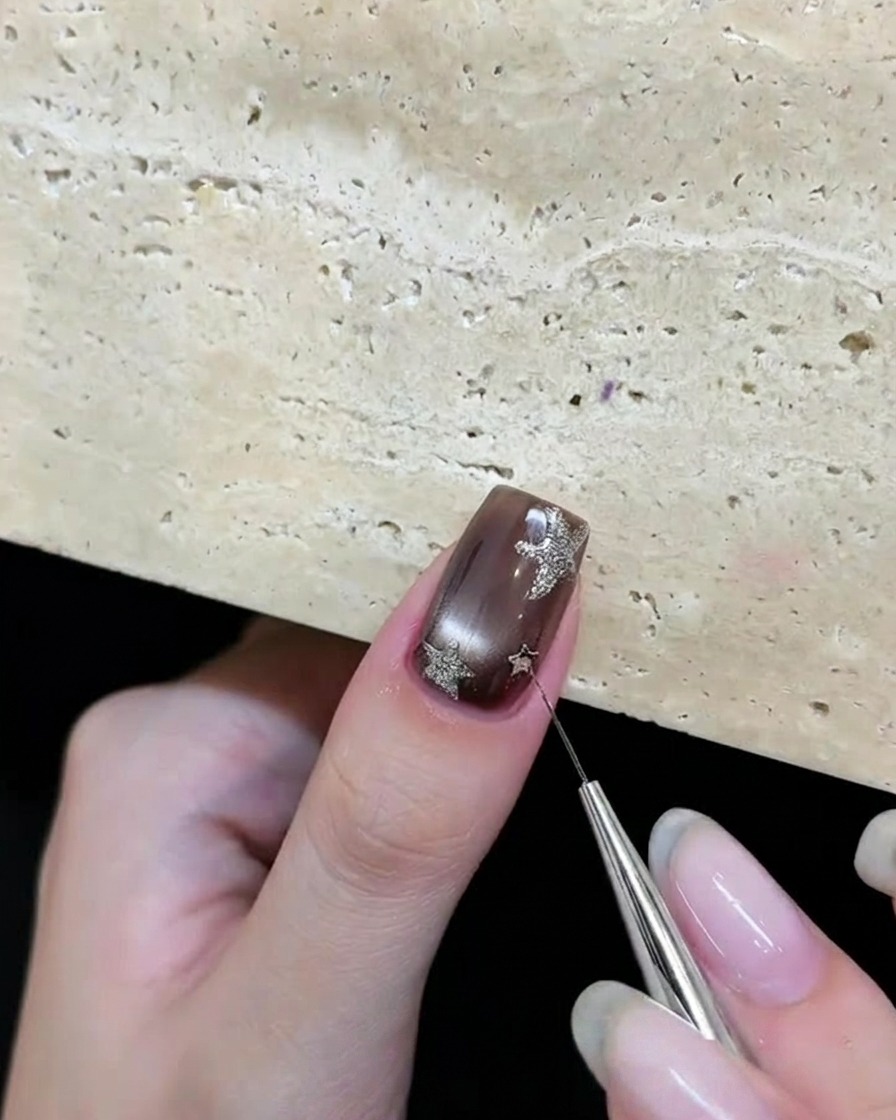 Uno strumento posiziona frammenti di glitter a forma di stella argentata su ununghia in gel marrone.