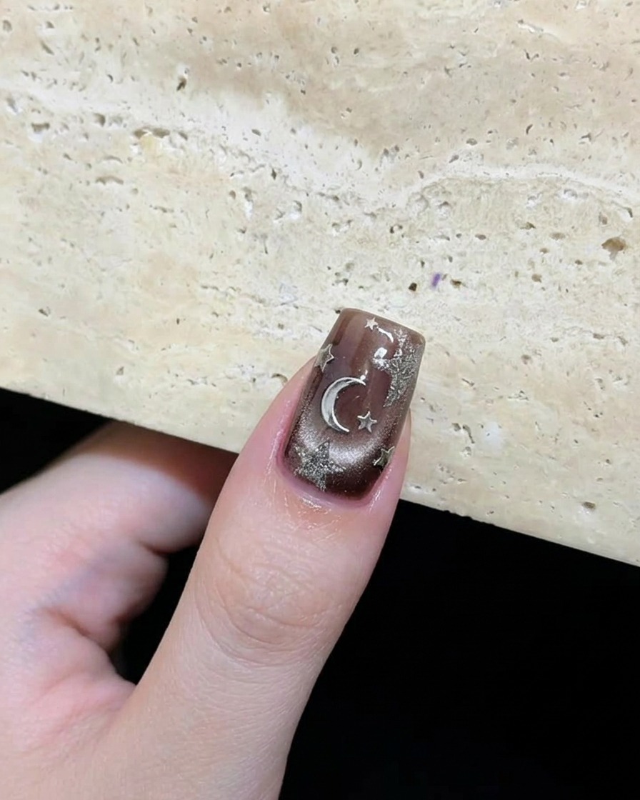 Primo piano di ununghia finita marrone scuro con stelle argentate e una luna decorativa disposte artisticamente.