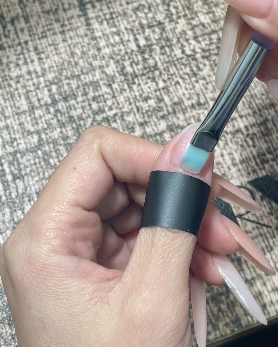 Primo piano di un pennellino che applica uno smalto gel nude traslucido su ununghia naturale nuda e preparata.