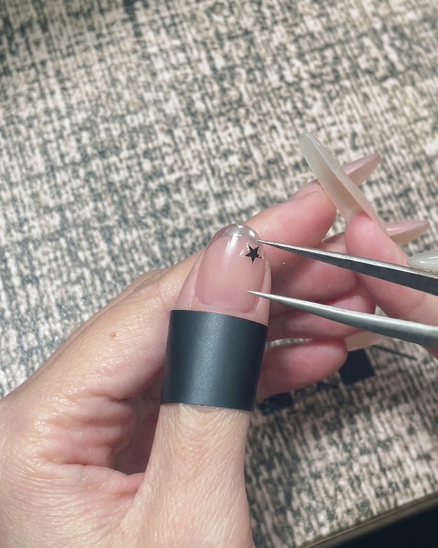 Una mano che tiene un paio di pinzette per posizionare un piccolo adesivo a forma di stella nera su ununghia in gel nude.