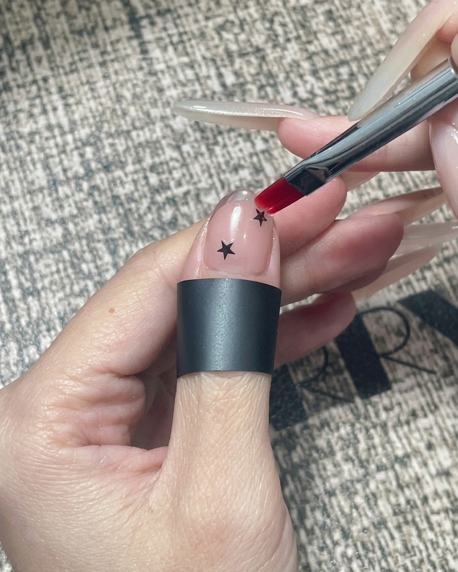Un pennello piatto per gel che applica gel trasparente incapsulante su ununghia decorata con stelle nere e una punta cromata.