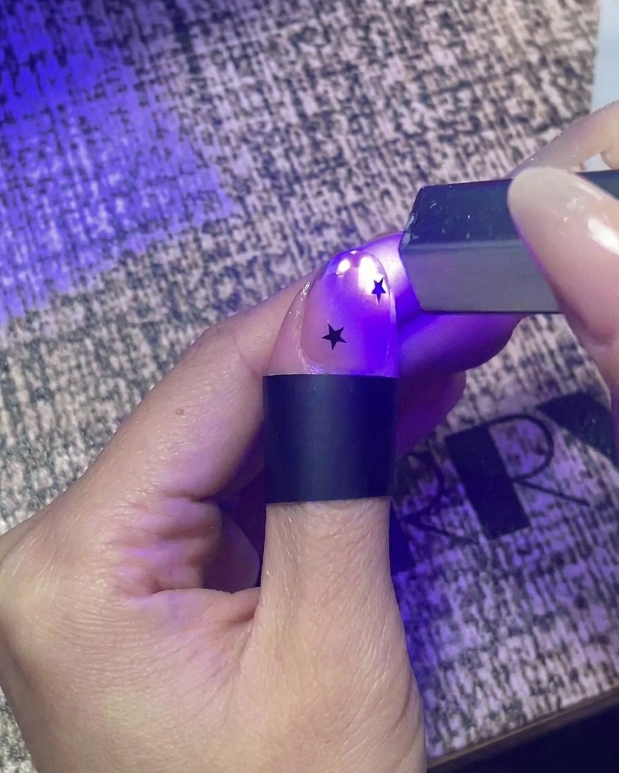 Primo piano di una mano che utilizza una piccola torcia UV portatile per polimerizzare rapidamente ununghia decorata con adesivi a forma di stella nera.