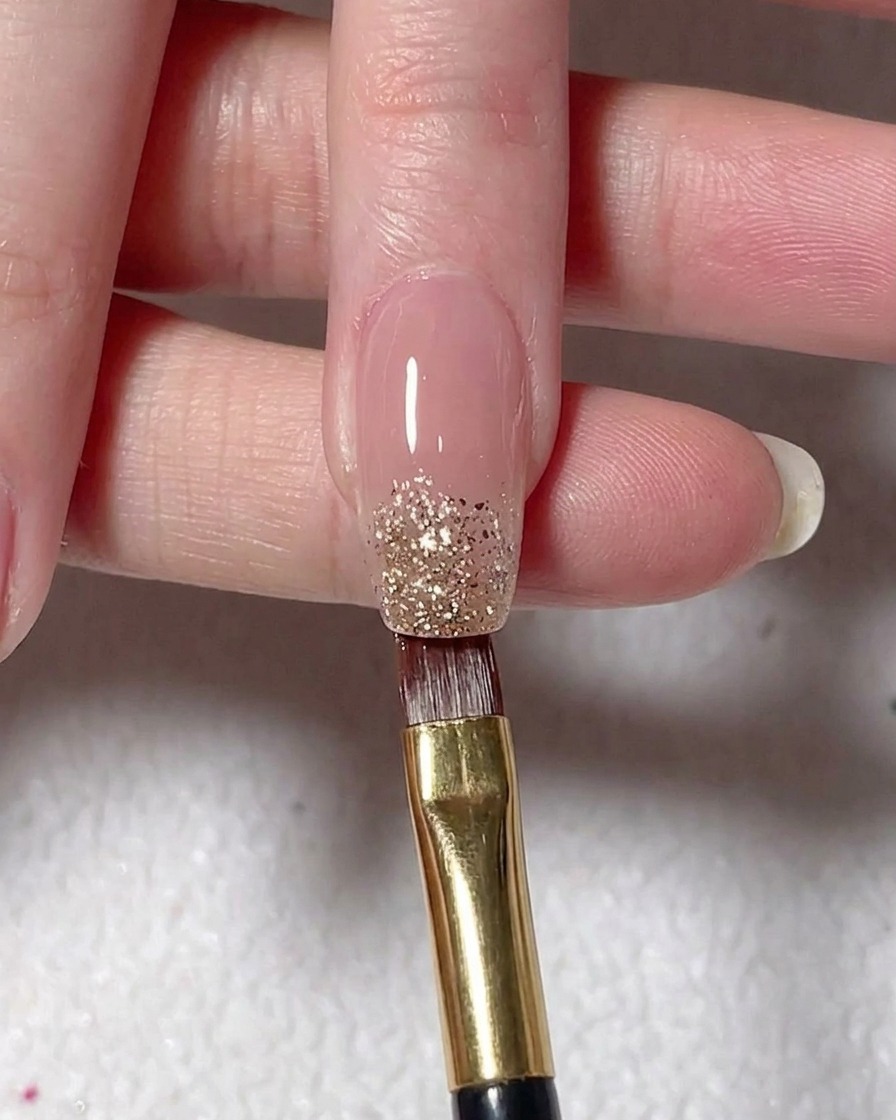 Un pennello piatto deposita gel glitter argento sulla punta di ununghia dipinta color nude.