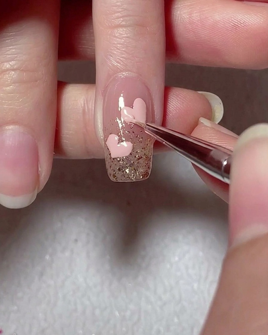 Un pennello liner rifinisce i bordi di un cuore rosa in gel su ununghia con accenti glitterati.