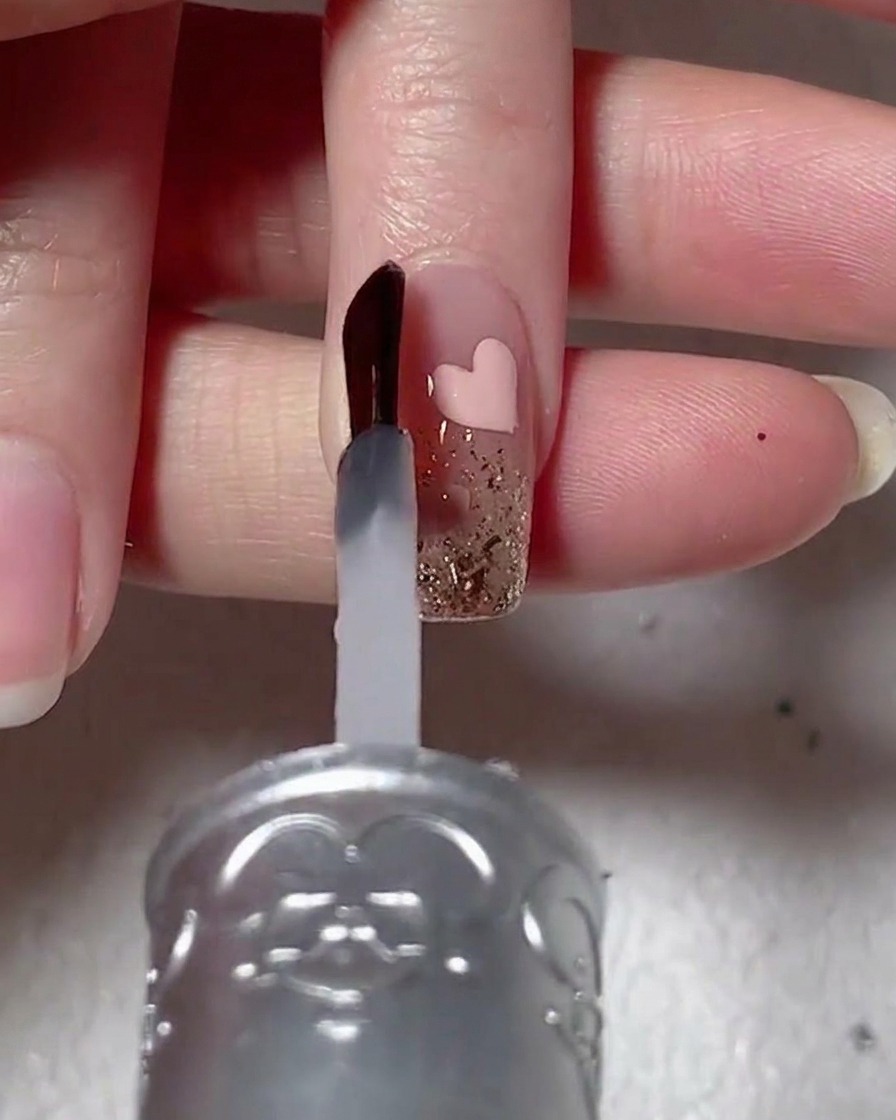 Un pennello per unghie applica un gel di rinforzo viscoso trasparente su ununghia decorata con glitter e un cuore rosa.