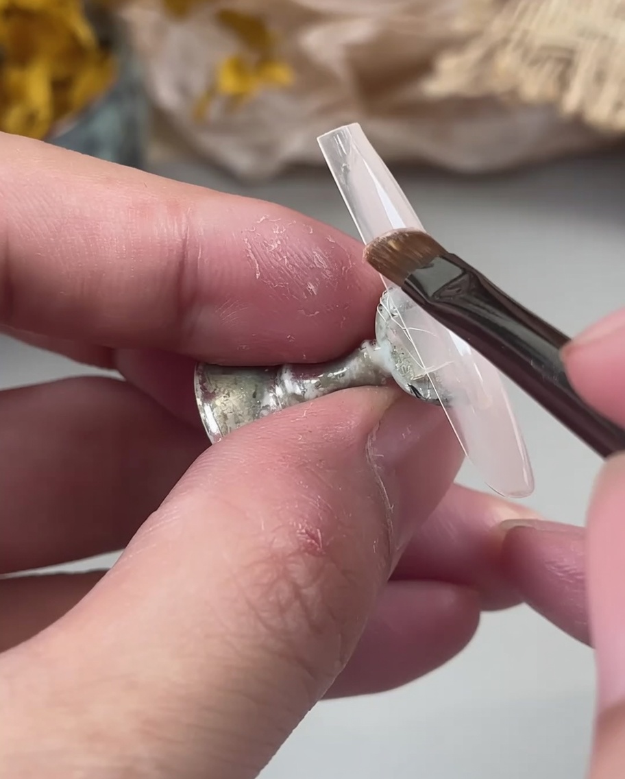 Un pennellino applica una base gel color nude lattiginoso su una tip trasparente tenuta tra le dita.